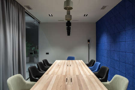 Conference room in a luminous office with gray and textured blue walls. There are wide wooden tables with multicolor chairs, glass partition with a door and curtain, green plants, different lamps.の写真素材