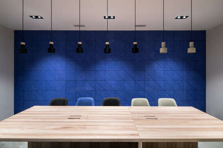 Conference room in a luminous office with gray and textured blue walls. There are wide wooden tables with multicolor chairs, hanging dark and light lamps. Horizontal.の写真素材