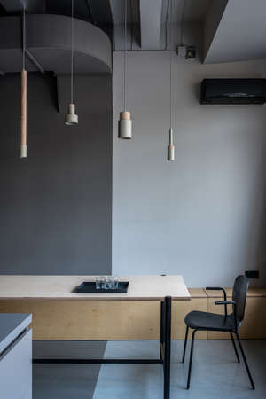 Nice room in an office in a loft style with gray walls. There is a wide wooden table with black tray with glasses, dark chair, different hanging beige metallic-wooden lamps, conditioner. Vertical.の写真素材