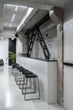 Stylish bar in a luminous interior in a loft style with gray walls and concrete columns. There is bar rack with a faucet and glass, wooden black partitions, dark chairs, armchair, green plant.の写真素材