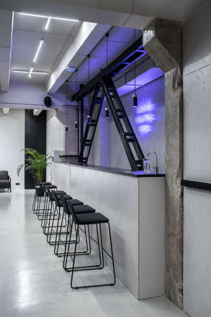 Trendy bar in a luminous interior in a loft style with gray walls and concrete columns. There is bar rack with a faucet and glass, wooden black partitions, dark chairs, armchair, green plant.の写真素材