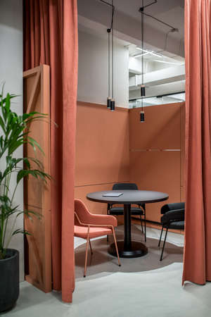 Zone for discussions with orange partitions and curtains in a luminous office in a loft style with gray walls. There is a round black table with a laptop, multicolor chairs, green plant in a big pot.の写真素材