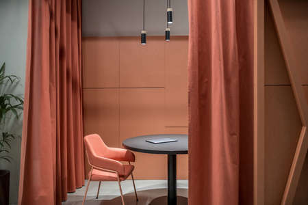 Place for discussions with orange partitions and curtains in an office in a loft style with gray walls. There is a round black table with a laptop, orange chair, green plant, luminous hanging lamps.の写真素材