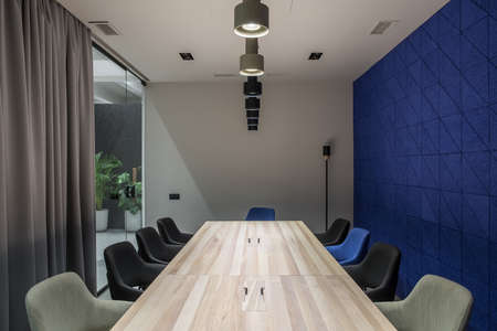 Stylish luminous office with a meeting room with gray and textured blue walls. There are wide wooden tables with multicolor chairs, glass partition with a door and curtain, plants, different lamps.の写真素材