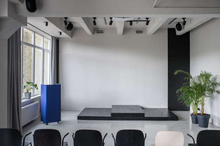 Great conference hall with gray walls and a glossy floor. There is a row of multicolor chairs, metallic blue stand, green plants in the pots, window with curtains, lamps and speakers. Horizontal.の写真素材