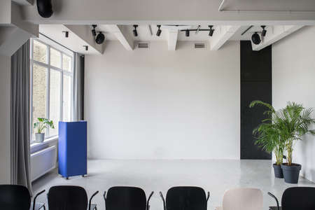 Empty conference hall with gray walls and a glossy floor. There is a row of multicolor chairs, metallic blue stand, green plants in the pots, window with curtains, lamps and speakers. Horizontal.の写真素材