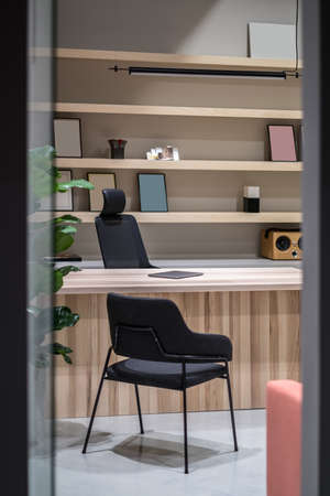 Room in a luminous office with gray walls. There is a wooden table with a laptop, black chairs, green plant, hanging lamp, wooden shelves with frames and a speaker and books. Vertical.の写真素材