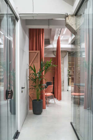 Hall in a luminous office in a loft style with gray walls and concrete columns. There are glass partitions with curtains, meeting zone with orange curtains, tables, chairs, green plant, shelves.の写真素材