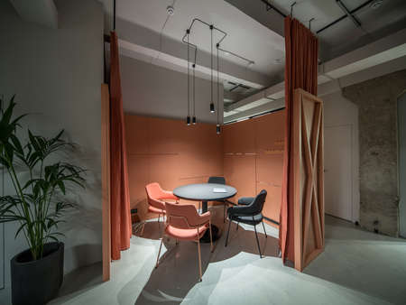Talk zone with orange curtains in a luminous office with gray walls and concrete column. There is a black round table with a laptop, multicolor chairs, partition, plant in a pot, hanging lamps.の写真素材