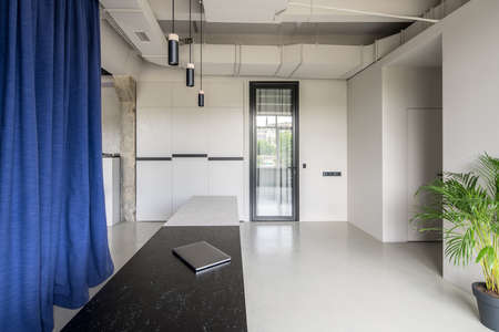 Nice office in a loft style with gray walls and a glossy floor. There is a dark-light textured reception desk with a laptop, curtains, hanging lamps, green plant in a pot, glass door, concrete column.の写真素材