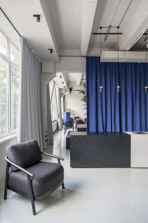 Stylish office in a loft style with gray walls and a glossy floor. There are multicolor chairs, dark-light textured reception desk, curtains, windows, black armchair, hanging lamps, green plants.の写真素材