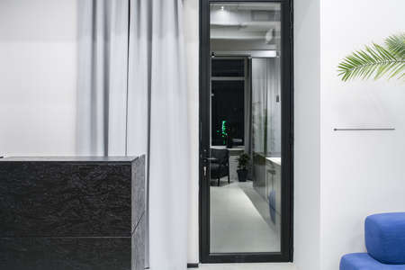 Textured black reception desk on the background of the gray wall with a glass door and curtain in an office. There is a blue sofa with a green plant on the right side. Horizontal.の写真素材