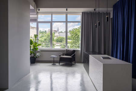 Interior in an office in a loft style with gray walls and a glossy floor. There is a textured black-white reception desk with a laptop, dark armchair, small round table, curtains, window, green plant.の写真素材