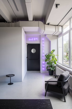Hall in an office in a loft style with gray walls and a glossy floor. There is a textured black reception desk, dark armchair, small round table with a laptop, green plant, door with a round window.の写真素材