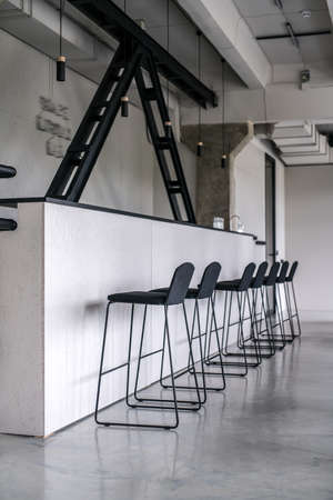 Stylish bar with a rack and black wooden partitions in an interior in a loft style with gray walls and concrete columns. There are several chairs, glasses and faucet, hanging lamps, glass door.の写真素材