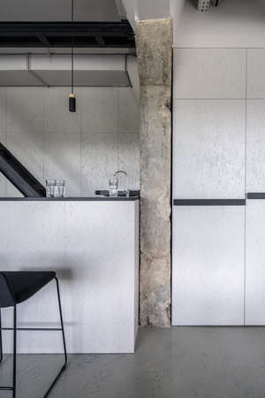 Beautiful bar with a rack and black wooden partition in an interior in a loft style with gray walls and a concrete column. There is a chair, glasses and faucet, hanging lamp, textured doors. Vertical.の写真素材