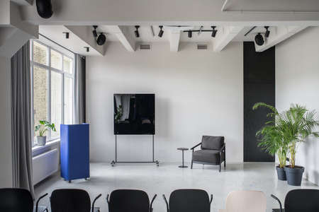 Light conference hall with gray walls and a glossy floor. There is a row of multicolor chairs, metallic blue stand,dark armchair, small round table, black board, green plants, window with curtains.の写真素材