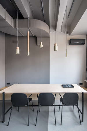Meeting room in an office in a loft style with gray walls. There is a wide wooden table with black tray with glasses, three dark chairs, different hanging beige metallic-wooden lamps, conditioner.の写真素材