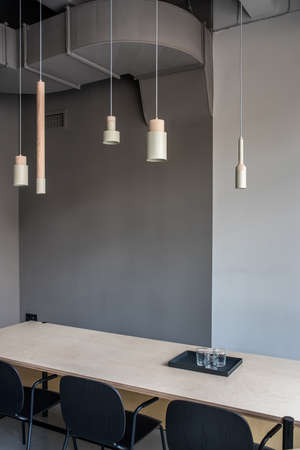 Stylish meeting room in an office in a loft style with gray walls. There is a wide wooden table with black tray with glasses, three dark chairs, different hanging beige metallic-wooden lamps.の写真素材