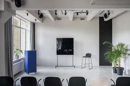 Beautiful conference hall with gray walls and a glossy floor. There are multicolor chairs, metallic blue stand, black board, green plants in the pots, window with curtains, lamps and speakers.の写真素材