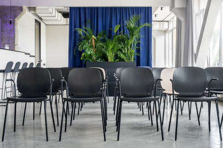 Nice conference hall in a loft style with gray walls and a concrete column. There are many multicolor chairs, bar, windows with curtains, black stand with green plants, hanging lamps, glass door.の写真素材