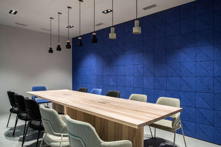 Meeting room with luminous lamps in an office with gray and textured blue walls. There are wide wooden tables with multicolor chairs, hanging dark and light lamps. Horizontal.の写真素材