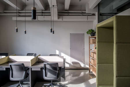 Room in an office in a loft style with gray walls. There are black tables with partitions, chairs, wooden shelves with boxes and a plant, green fancy armchair, board, hanging lamps. Horizontal.の写真素材