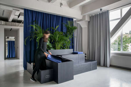 Room in an office in a loft style with gray walls and a glossy floor. There are wooden black stands with lockers and green plants and blue pillows, windows, multicolor curtains, doors, mirror, woman.の写真素材