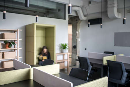 Room in a loft style with gray walls. There are black tables with multicolored partitions, chairs, wooden shelves with boxes and plants, green fancy armchair with a woman with a book, glass door.の写真素材