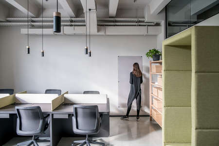 Office in a loft style with gray walls. There are black tables with partitions, chairs, wooden shelves with boxes and a plant, green fancy armchair, board, hanging lamps, woman. Horizontal.の写真素材