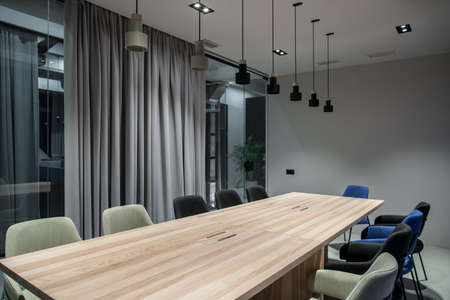 Meeting room with a luminous lamps in an office with gray walls. There are wide wooden tables with multicolor chairs, glass partition with a door and curtain, different lamps, green plants.の写真素材