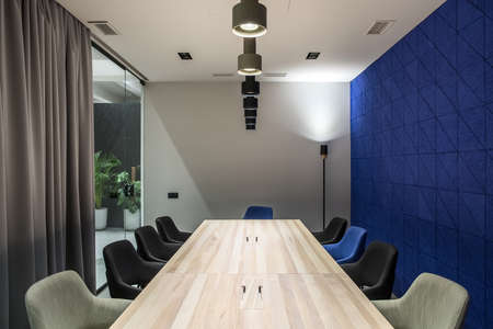 Discussion room in a luminous office with gray and textured blue walls. There are wide wooden tables with multicolor chairs, glass partition with a door and curtain, green plants, different lamps.の写真素材