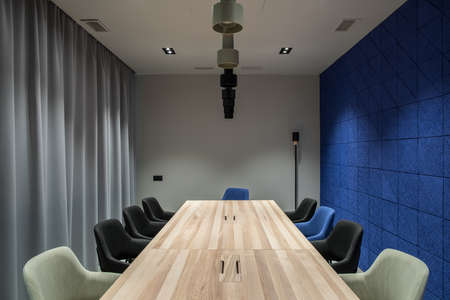 Meeting room with a luminous lamps in an office with gray and textured blue walls. There are wide wooden tables with multicolor chairs, curtains, different lamps. Horizontal.の写真素材