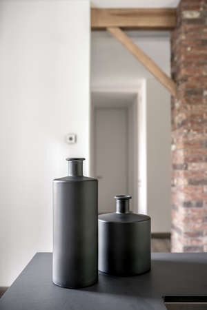 Two black vases on the dark tabletop on the background of the modern interior with white and brick walls and wooden beams. Low aperture closeup photo. Vertical.の写真素材