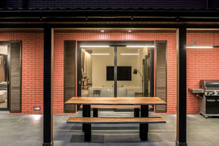 Fancy modern country house with luminous lamps. It has large glass doors, awning, terrace with a tiled floor and a wooden table with benches, BBQ. Inside there is a sofa with a TV and other furniture.の写真素材