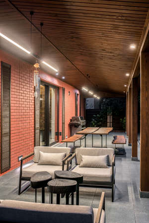 Stylish modern country house with luminous lamps and a wooden awning. It has a terrace with a tiled gray floor and tables with benches and armchairs with pillows, BBQ, fence. Vertical.の写真素材