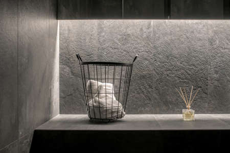 Illuminated modern bathroom with tiled textured gray walls. There a metal black basket with a white towel, aroma sticks, mirror. Closeup horizontal photo.の写真素材