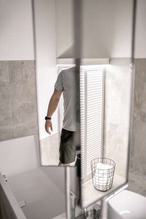 Contemporary bathroom with illumination, white and tiled gray walls. There is a bath, white sink, chrome faucets, mirrors, black basket with a towel, standing person. Vertical.の写真素材