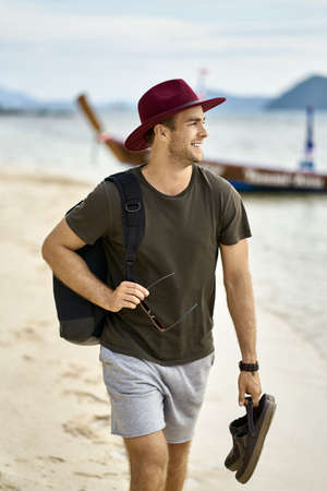 Barefoot man with stubble and backpack is walking on sand beachの写真素材