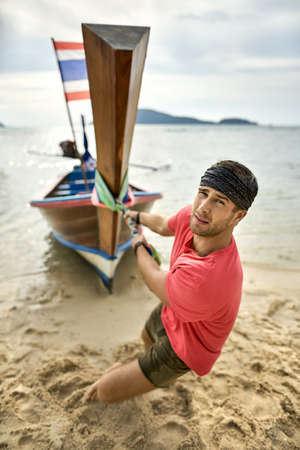 Man with stubble is pulling wooden boat by rope on sand beachの写真素材