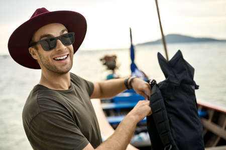 Cheerful man with stubble is holding his backpack near wooden boatの写真素材