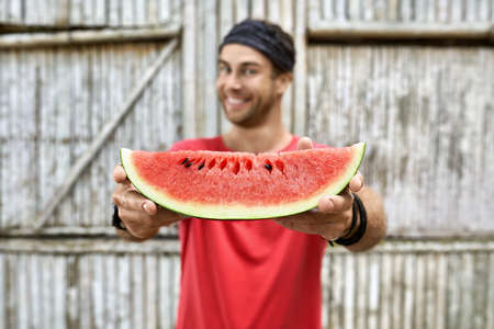 Smiling man with stubble holds slice of watermelon on wooden fence backgroundの写真素材