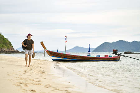 Barefoot man with stubble and backpack is walking on sand beachの写真素材