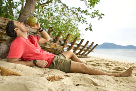 Man with stubble is drinking from coconut on sand beachの写真素材