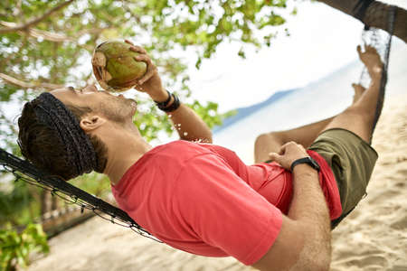 Man with stubble is drinking from coconut on hammock on sand beachの写真素材