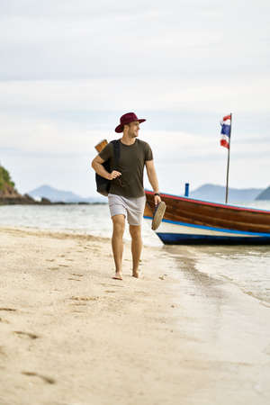 Barefoot man with stubble and backpack is walking on sand beachの写真素材
