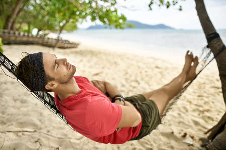 Man with stubble is lying on black hammock on sand beachの写真素材
