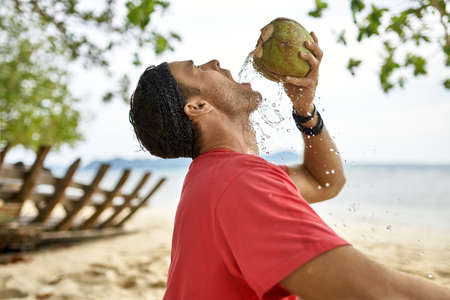 Man with stubble is drinking from coconut on sand beachの写真素材
