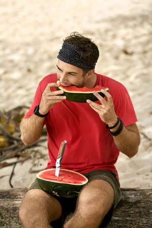Man with stubble is eating watermelon on sand beachの写真素材