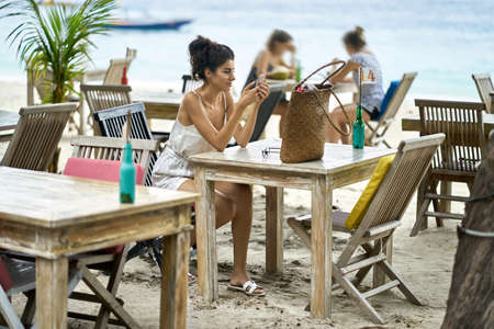 Tanned woman using her smartphone in outdoor restaurant on tropical beachの写真素材